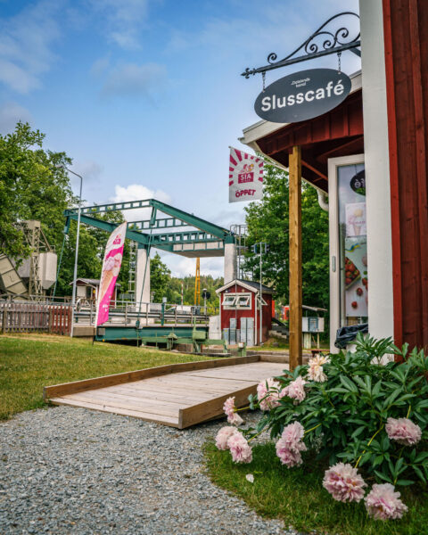 Dalslands kanals Slusscafé i Långbron, en röd stuga med skylt, rosa pioner, träramp och slussanläggningen i bakgrunden en sommardag.