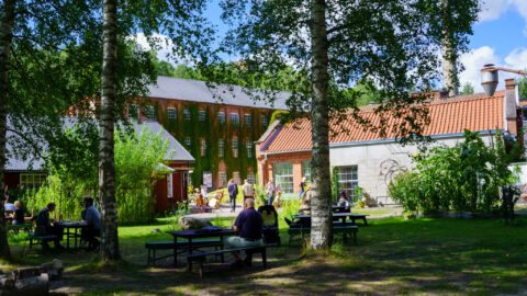 Uteservering med bänkbord bland björkar vid Not Quite Café i Fengersfors, med den gamla bruksbyggnaden i bakgrunden en solig sommardag