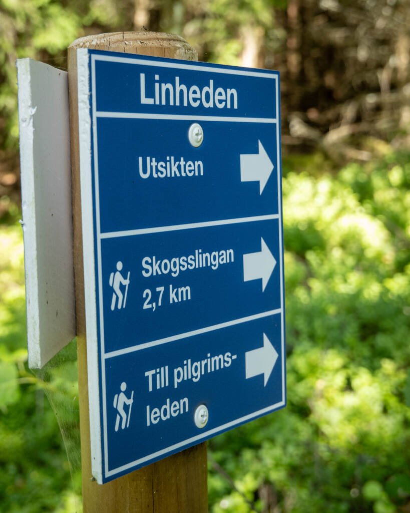 Blå vägvisarskylt vid Linheden i Dalsland med pilar mot Utsikten, Skogsslingan 2,7 km och Pilgrimsleden, omgiven av grön skog.