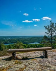 Träbänk på berghäll vid Linhedens utsiktsplats i Dalsland med vidsträckt vy över sjöar och grön skog under blå himmel