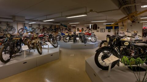 Utställningshall i Eds MC- och Motormuseum med veteranmotorcyklar på podier, cyklar längs väggarna och svenska flaggor i bakgrunden