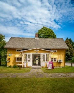 Gulmålat sommarcafé med gulvitrandiga markiser i Dals Rostock, en kvinna står i dörröppningen till Örtagårdens Kafé omgivet av grönska.