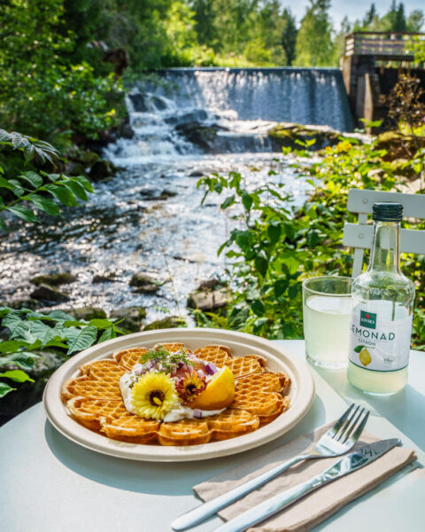 Svensk våffla med grädde och blommor serverad med Kiviks citronlemonad vid Tollebols Kvarncafé, med forsande vatten och grönska i bakgrunden.