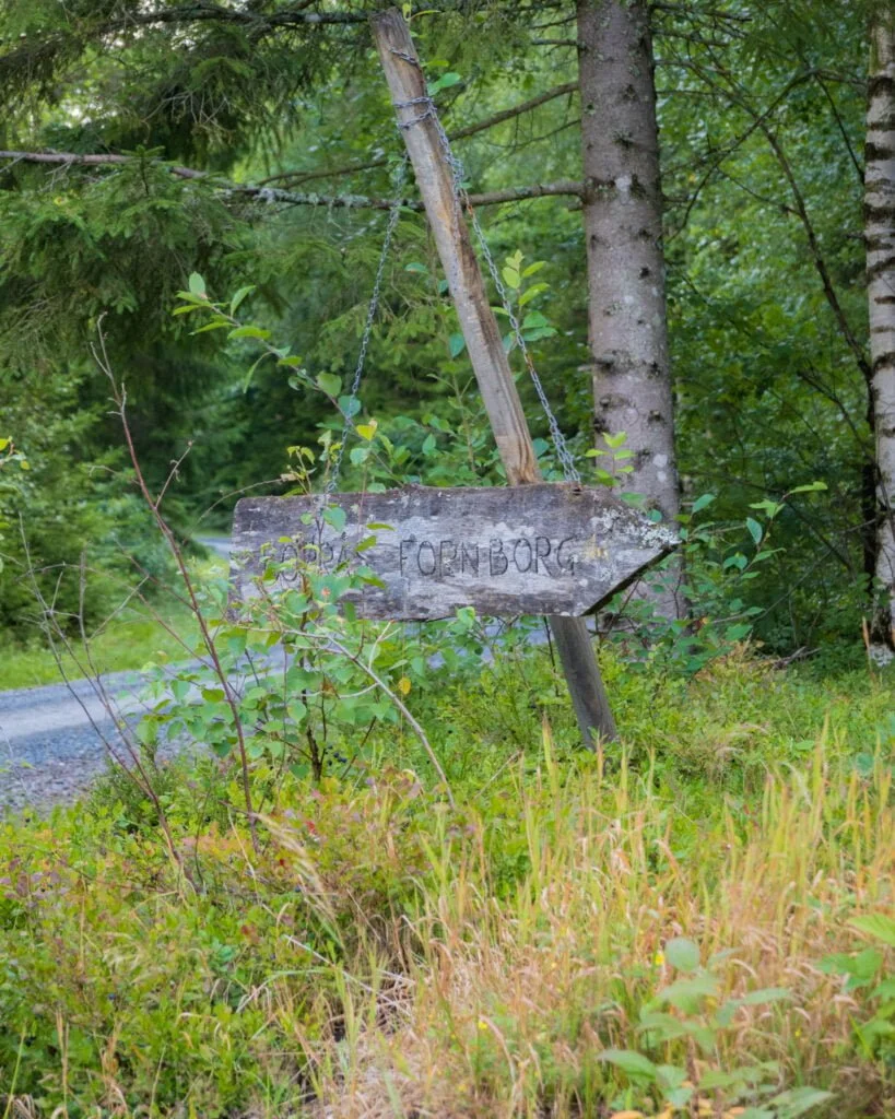 Vädersliten träskylt med texten "Fornborg" hängande i kedjor vid en skogsväg nära Borråsbergets fornborg i Färgelanda, Dalsland