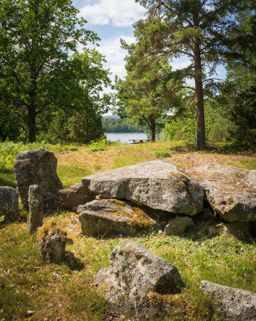 Forntida hällkista med stora stenhällar på en gräsbeklädd höjd omgiven av lövträd och tallar, med utsikt över sjön Ånimmen i Dalsland