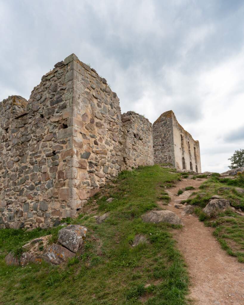 Brahehus ruin med massiva stenmurar och bågfönster på en gräsbevuxen kulle, en stig leder upp mot 1600-talsruinen i Småland.