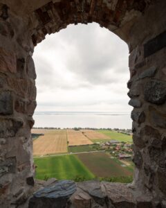 Utsikt över jordbrukslandskap och Vättern genom ett bågformat stenfönster i Brahehus ruin, Småland