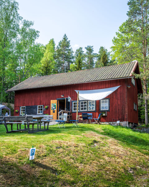 Röd träbyggnad med solsegel och utemöbler på grösmatta vid Nyfiket Ektorp, ett sommarcafé och gårdsbutik utanför Finspång, omgivet av lövträd.