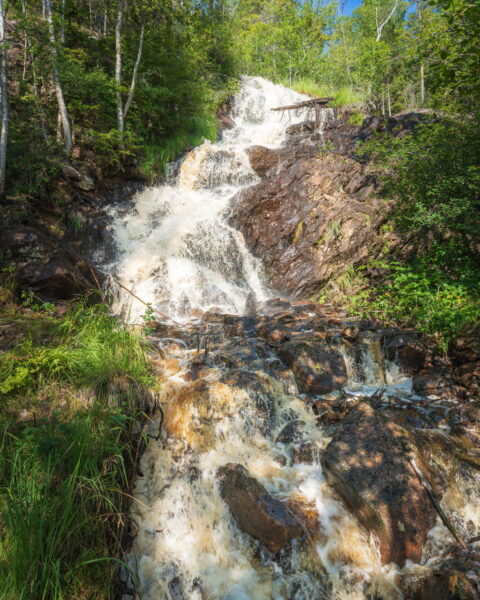 Vattenfallet Brudslöjan utanför Dals-Ed i Dalsland, med forsande vatten över röda klippor omgivet av grön björkskog en sommardag