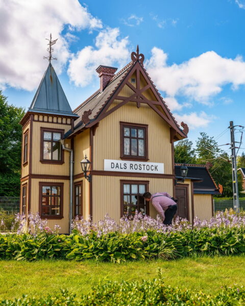 Dals-Rostocks mini-stationshus i skala 1:2, en gulbeige träbyggnad med bruna detaljer och torn, omgiven av blommande rabatter och grön gräsmatta.