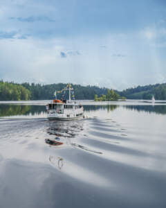 Vit passagerarbåt med svenska flaggor glider fram på spegelblank vattenyta i Dalslands kanal, omgiven av grönskande skogsklädda stränder.
