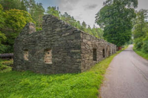 Stenruiner av Bäckefors gamla järnbruk i Dalsland, med murade väggar och fönsteröppningar längs en smal asfalterad stig omgiven av grönska.