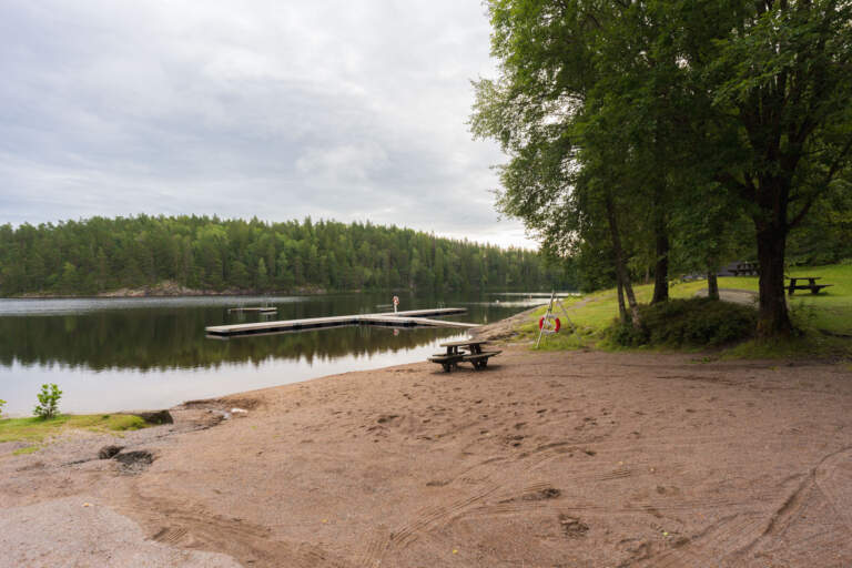 Sandstrand vid Sjöluckan i Bäckefors med badbrygga, bänk och livboj vid en stilla sjö omgiven av grön barrskog i Dalsland.