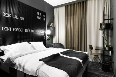 Modernt hotellrum på The Box i Linköping med dubbelsäng, svartvit inredning och pegboard-vägg med vit text om priser och hashtags.