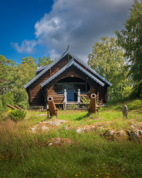 Ödeborgs fornsal, en karaktäristisk timmerstuga med vikinginspirerat spetsigt tak, omgiven av grönska och gamla kanoner i Färgelanda kommun, Dalsland