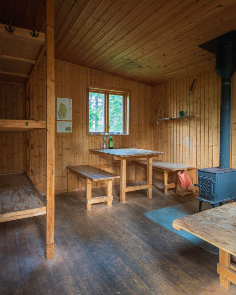 Interiör i raststugan på Baljåsen med träbord, bänkar, vedspis och britsar i en ombonad stuga med träpanel och fönster mot skogen