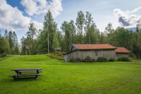 Petersborg hembygdsgård i Baljåsen med grått timmerhus, tegeltak, picknickmöbler på grön gräsmatta, klockstapel och svensk flaggstång omgivna av björkar.
