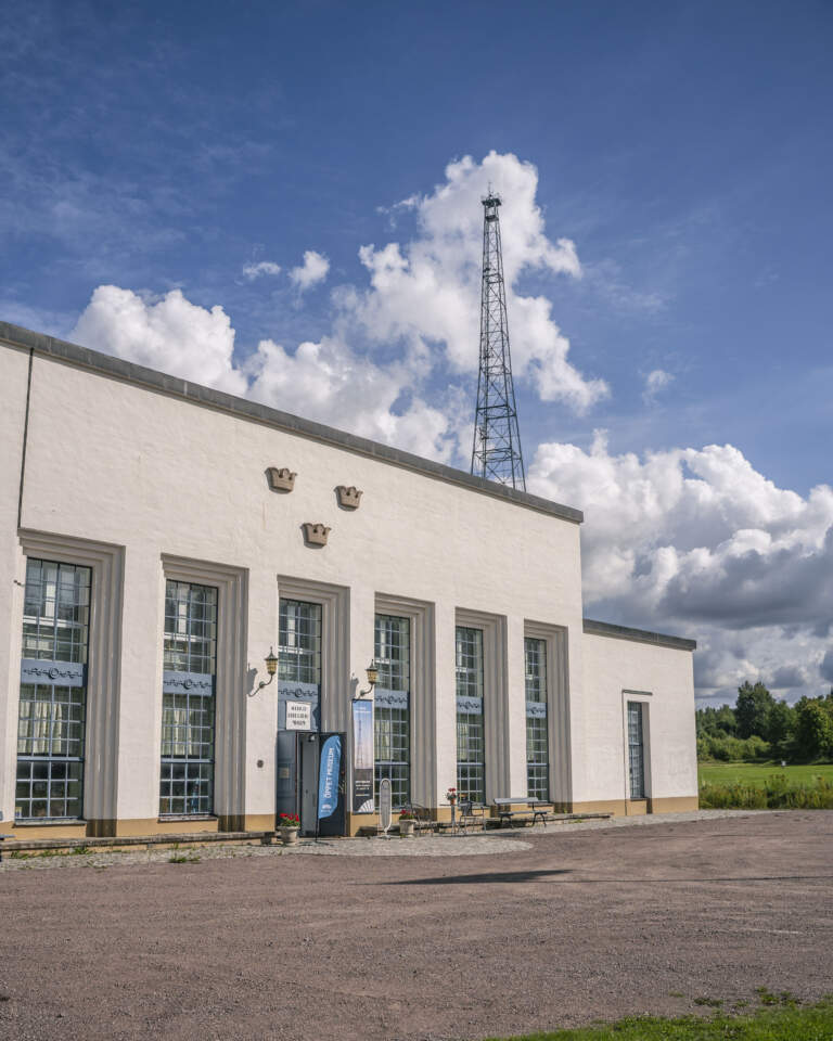 Sveriges Rundradiomuseum i Motala, en vitputsad byggnad med tre kronor på fasaden och en radiomast i bakgrunden, en solig sommardag