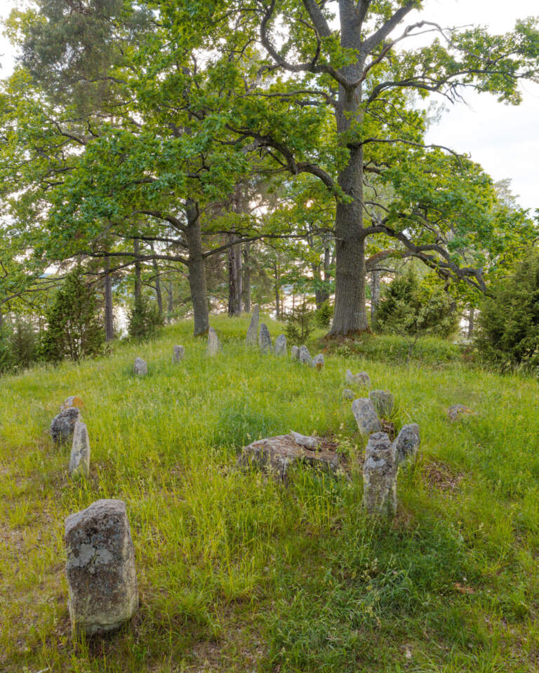 Skeppssättning på Gumhems gravfält vid Jätteudden, med resta stenar i grönt gräs under stora lövträd vid sjön Åsunden