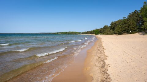 Varamobadets långa sandstrand vid Vättern i Motala med turkost vatten, små vågor och tallskog längs strandkanten en solig dag