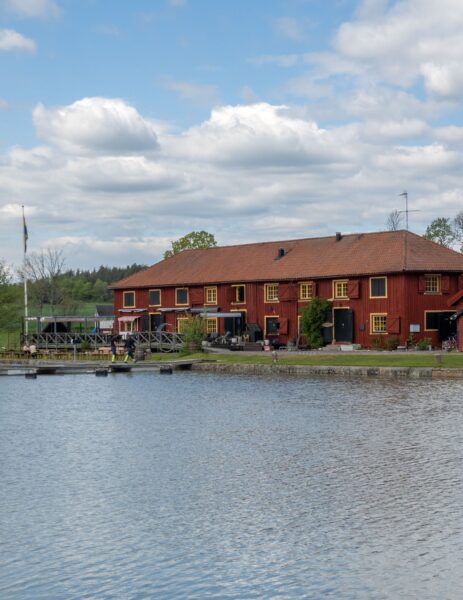 Kanalmagasinet i Mem, en faluröd byggnad med gula fönster vid vattnet där Göta kanal möter Slätbaken, med brygga och flaggstång.