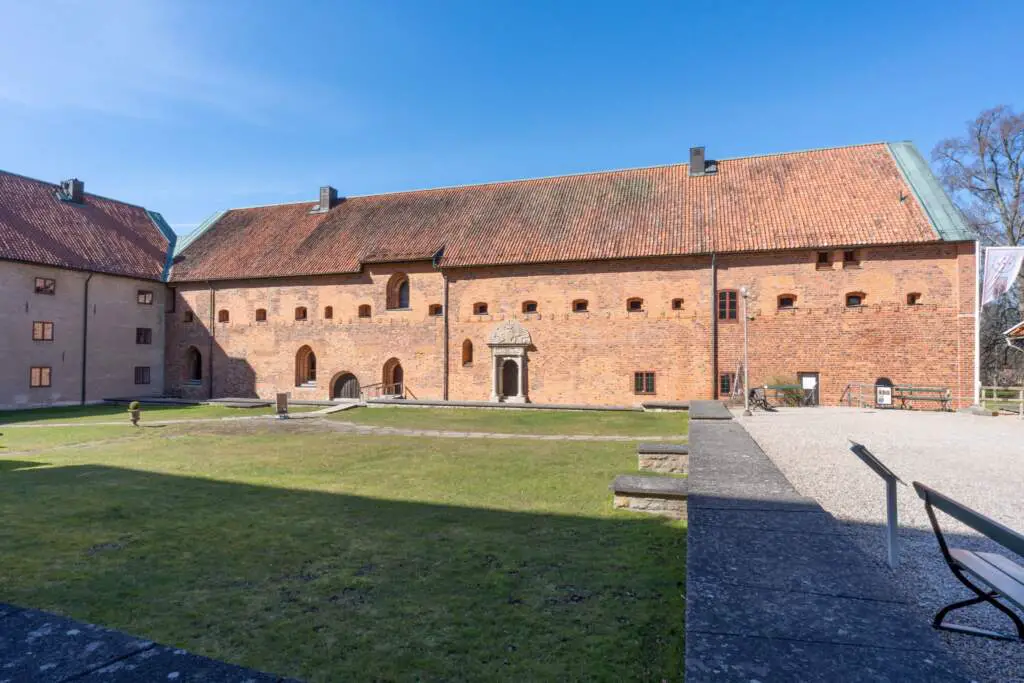 En solig vy av Sancta Birgitta Klostermuseum, som visar tegelstensbyggnader med tak av rött tegel. En grön gräsmatta sträcker sig framför byggnaden, och det finns en liten stentrappa som leder till en trädörr. I förgrunden finns en grusgång, bänkar och ett stenmonument.