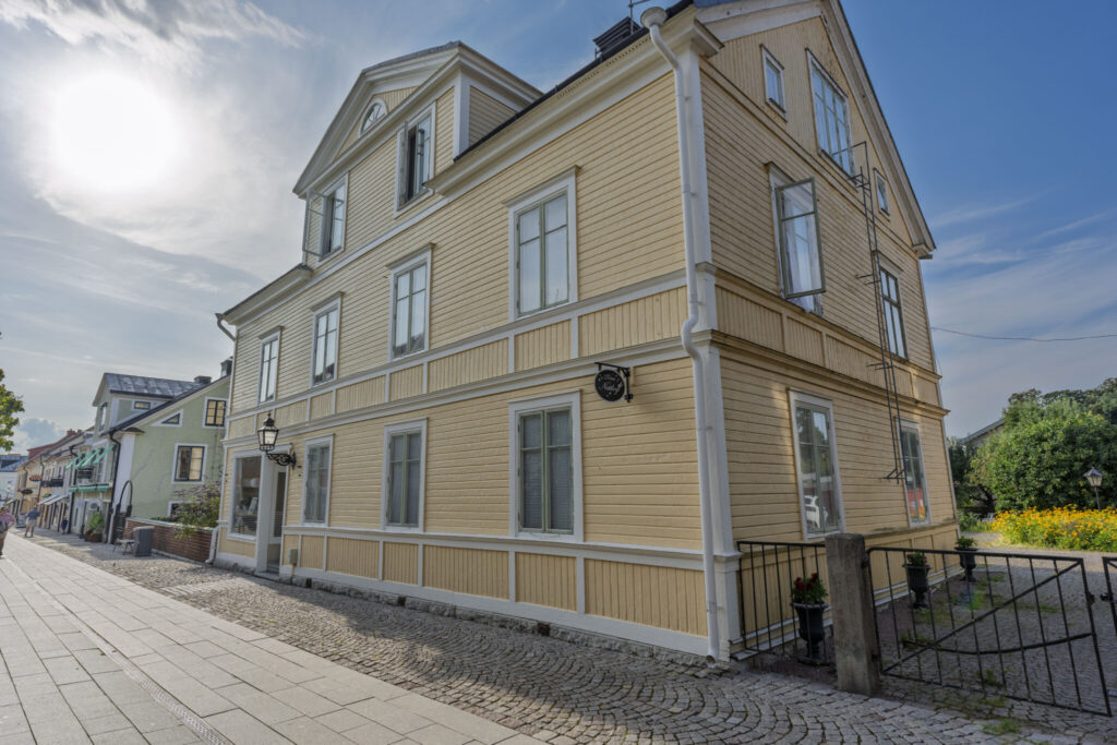 Gulmålat trähus med vita detaljer och kullersten framför 27ans Nattlogi i centrala Vadstena, en charmig småstadsgata en sommardag