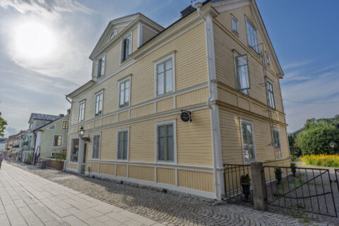 Gulmålat trähus med vita detaljer och kullersten framför 27ans Nattlogi i centrala Vadstena, en charmig småstadsgata en sommardag