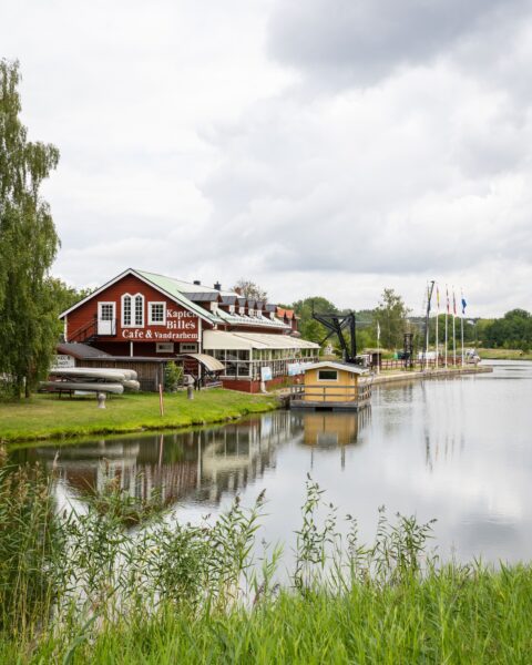 Kapten Billes Café & Vandrarhem, en röd träbyggnad vid Göta kanal i Norsholm, med brygga, flaggstänger och spegelblank vattenyta.