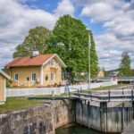 Norsholms slusscafé vid Göta kanal, gult trähus med vitt staket intill en slussport, omgivet av gröna lövträd en sommardag
