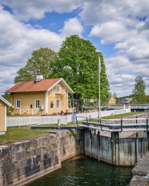 Norsholms slusscafé vid Göta kanal, gult trähus med vitt staket intill en slussport, omgivet av gröna lövträd en sommardag