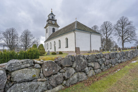 Rystads kyrka med vitputsad fasad, klocktorn och kyrkogård omgiven av en gammal stenmur en mulen höstdag i Östergötland