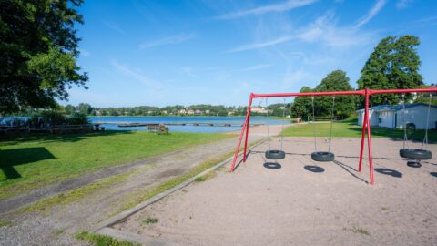 Röda däckgungor på sandplan vid Lilla Le's badplats i Ed, med gröna gräsytor, badbryggor och lugnt sjövatten en solig sommardag.