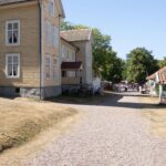 Gul träbyggnad längs grusgång i Medevi brunns historiska brunnsmiljö en solig sommardag, med besökare och marknadsstånd i bakgrunden