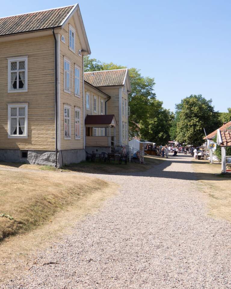 Gul träbyggnad längs grusgång i Medevi brunns historiska brunnsmiljö en solig sommardag, med besökare och marknadsstånd i bakgrunden