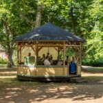 Brunnsorkestern spelar i en musikkiosk omgiven av lummiga lövträd i Medevi brunn en solig sommardag