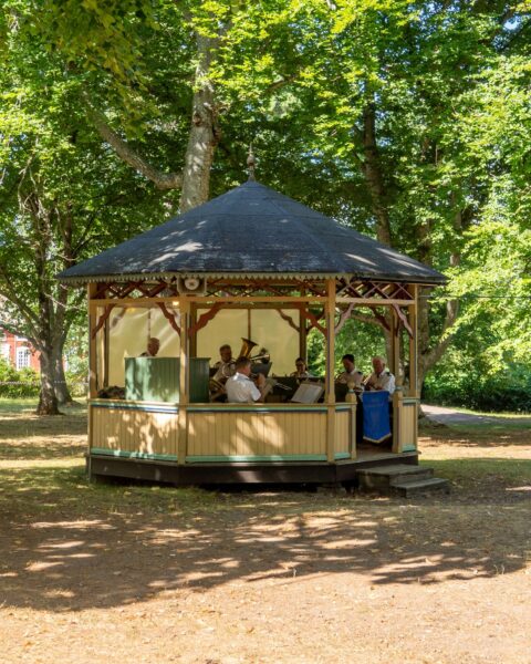 Brunnsorkestern spelar i en musikkiosk omgiven av lummiga lövträd i Medevi brunn en solig sommardag