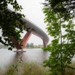 Modern bågbro med röda stöd sedd från Rödbergsudden i Motala, inramad av gröna lövträd vid vattnets kant en disig dag.