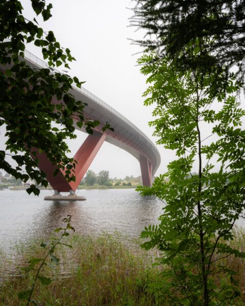 Modern bågbro med röda stöd sedd från Rödbergsudden i Motala, inramad av gröna lövträd vid vattnets kant en disig dag.