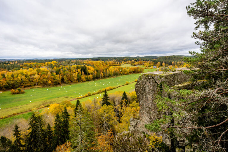 Höstvy från Klevbergets utsiktsplats i Kinda med branta klippor, gröna ängar med ensilabebalar och skog i höstfärger.