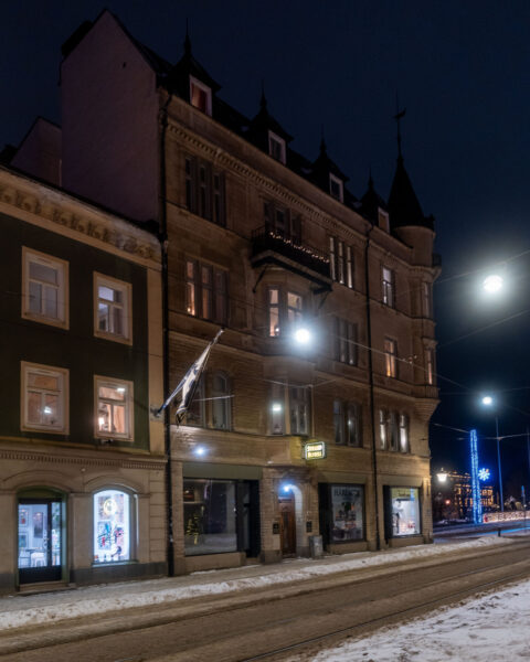 Strand Hotel i Norrköping en vinterkväll, ett charmigt 1800-talshus med upplysta butiksfönster, snö på gatan och julbelysning i bakgrunden