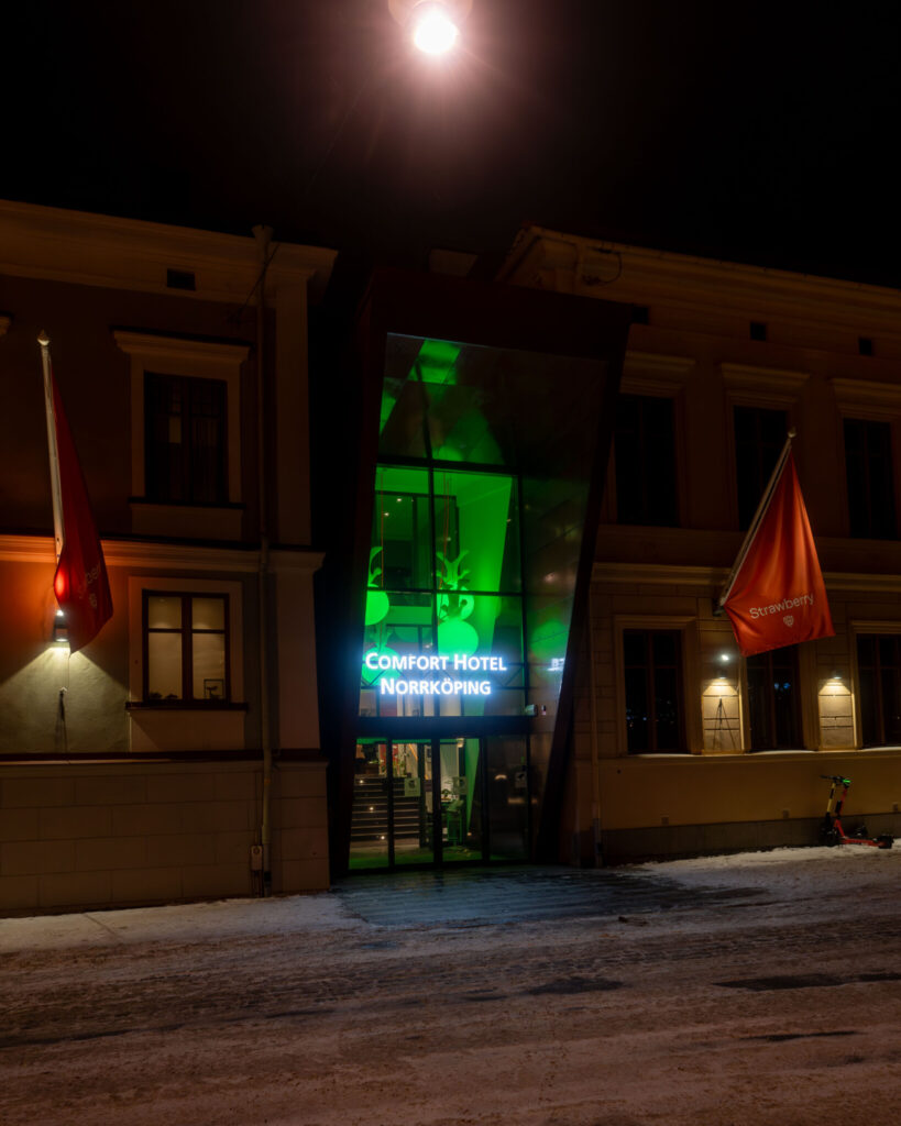 Comfort Hotel Norrköpings entré en vinterkvall med grönbelyst glasfasad, flaggor och snö på marken under en lysande måne.