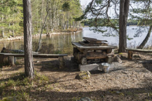 Grillplats med träbänkar vid Hämtningsviken i Västanvik, omgiven av tallskog med utsikt över stilla vatten en solig dag.