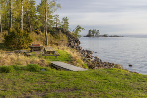 Grillplats vid Kagenviken i Västanvik med picknickbord, brygga och stenig strandkant mot stilla vatten och en liten skogsklädd ö.