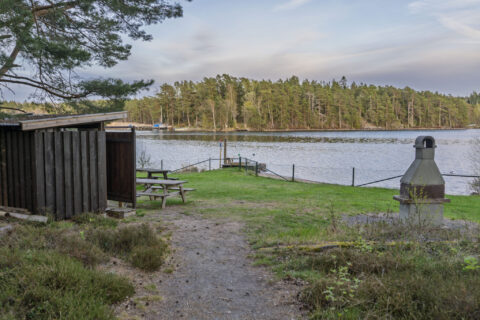 Grillplats vid Badudden i Västanvik med vindskydd, bänkbord och murad grill på en grön gräsyta vid sjön, omgiven av tallskog.