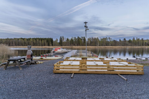 Grillplats vid Yttre Odensberg i Västanvik med kolgrill, bänkbord och småbåtsbrygga vid en stilla sjö omgiven av skog och vass.