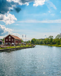 Röd kanalbyggnad vid Göta kanal i Borensberg med flaggstänger, grönskande träd och glittrande vatten en sommardag.
