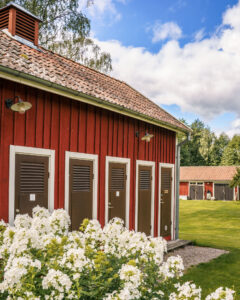 Röd servicebyggnad med bruna dörrar och vita karmar vid Glasbruket i Borensberg, omgiven av vita blommor och grön gräsmatta.