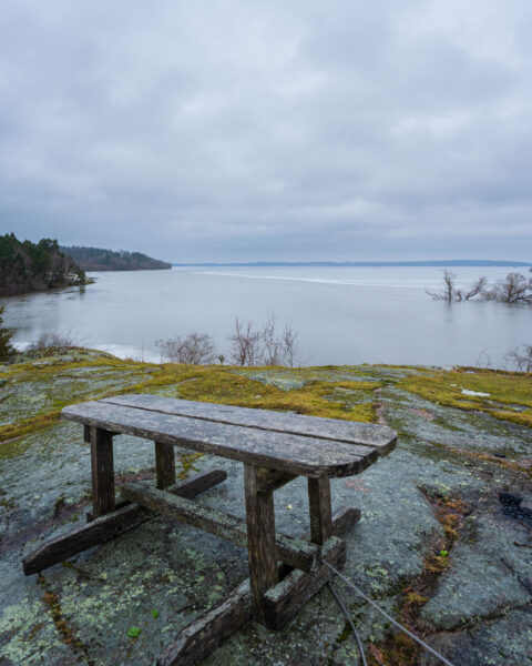 Väderbitен träbänk på mossklädd klipphäll vid Kungsbacken i Ljusfors naturreservat med utsikt över Glan en mulen vinterdag