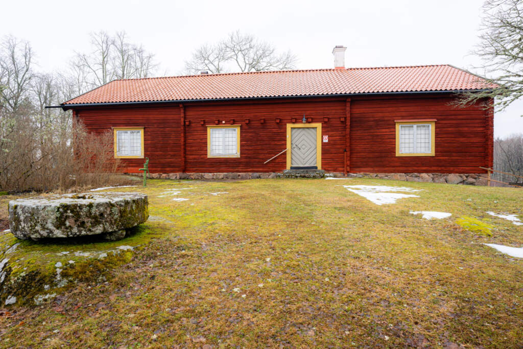 Faluröd timmerstuga vid Risinge Hembygdsmuseum i Finspång med gulmarkerade fönster, tegeltak och en stor stenkvarn på gräsmattan framför.